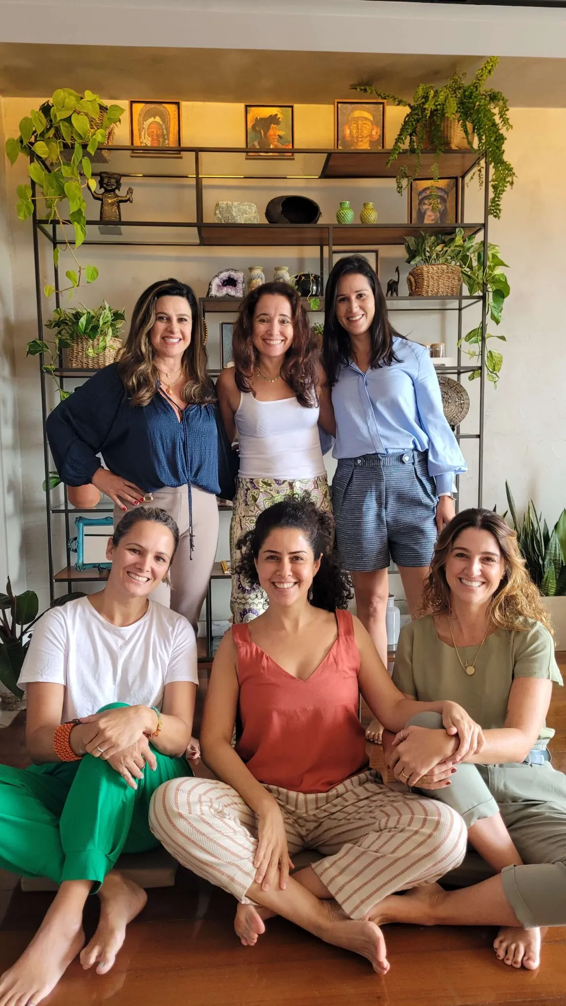 Grupo de sete mulheres reunidas em uma atividade presencial, sentadas e em pé diante de uma estante com plantas